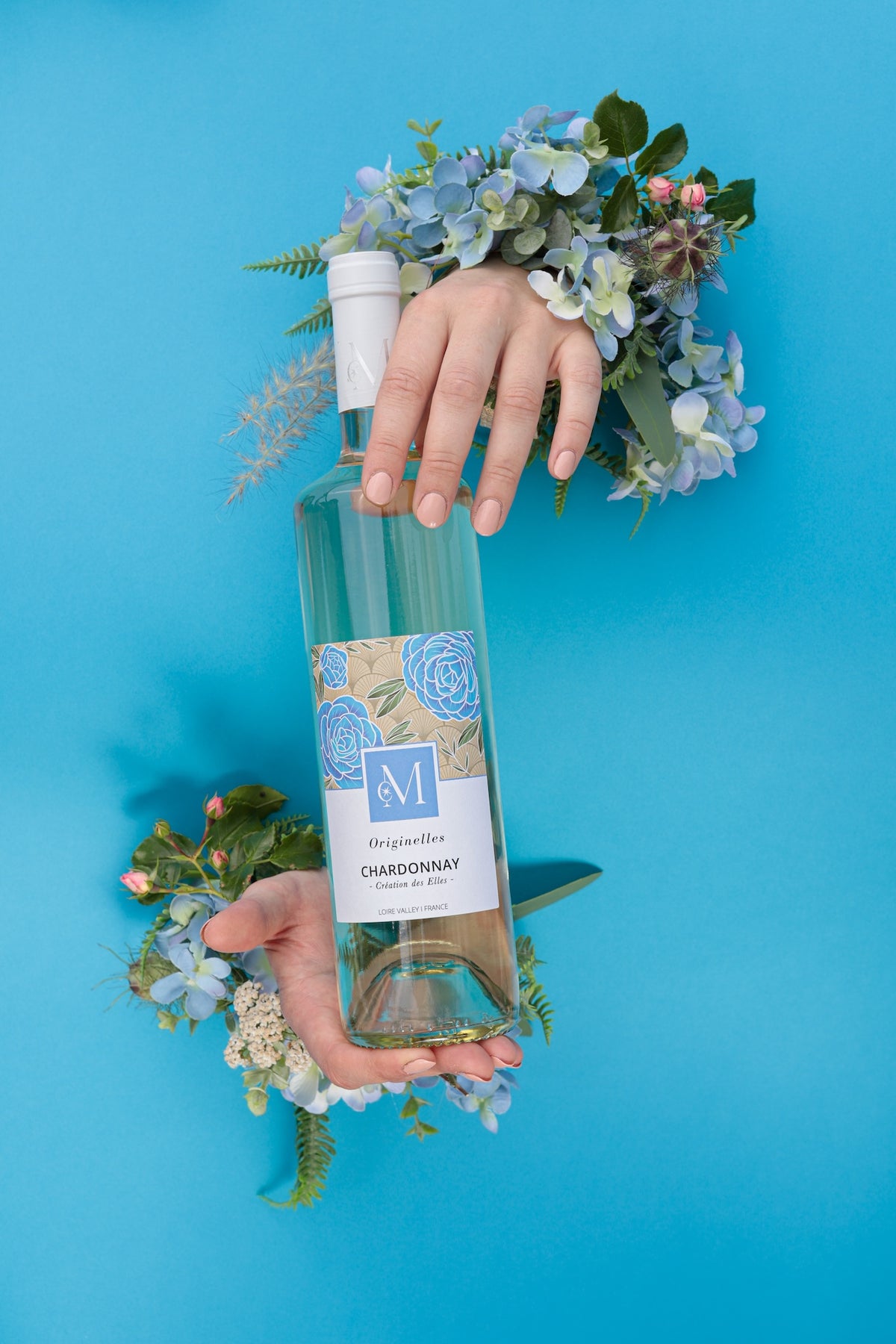 Bouteille de vin blanc Originelles Chardonnay du Moulin de Chauvigné tenue par deux mains entourées de fleurs, sur fond bleu