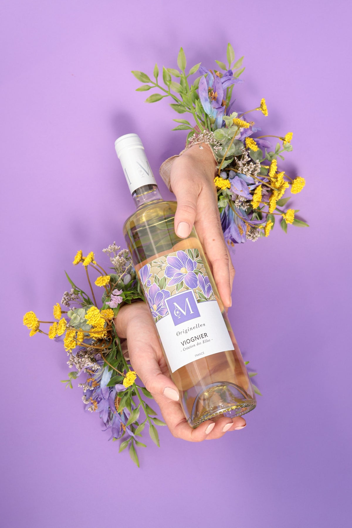 Bouteille de vin blanc Originelles Viognier du Moulin de Chauvigné tenue par deux mains entourées de fleurs violettes et jaunes, sur fond violet